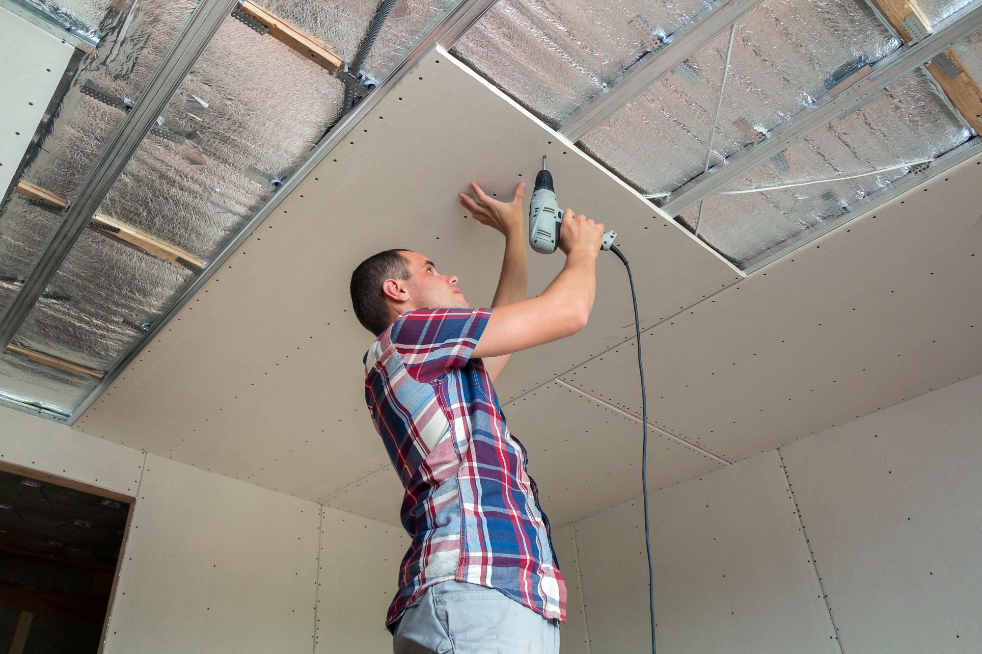 Young man in usual clothing and work gloves fixing drywall suspended ceiling